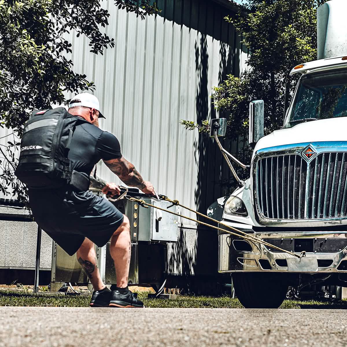 A muscular individual, outfitted in GORUCK's Men's Ballistic Trainers - HULK and athletic gear, pulls a large truck using ropes while wearing a weighted vest. The scene, highlighting the functional fitness qualities of this footwear, unfolds outdoors beside a metal building with trees in the background.