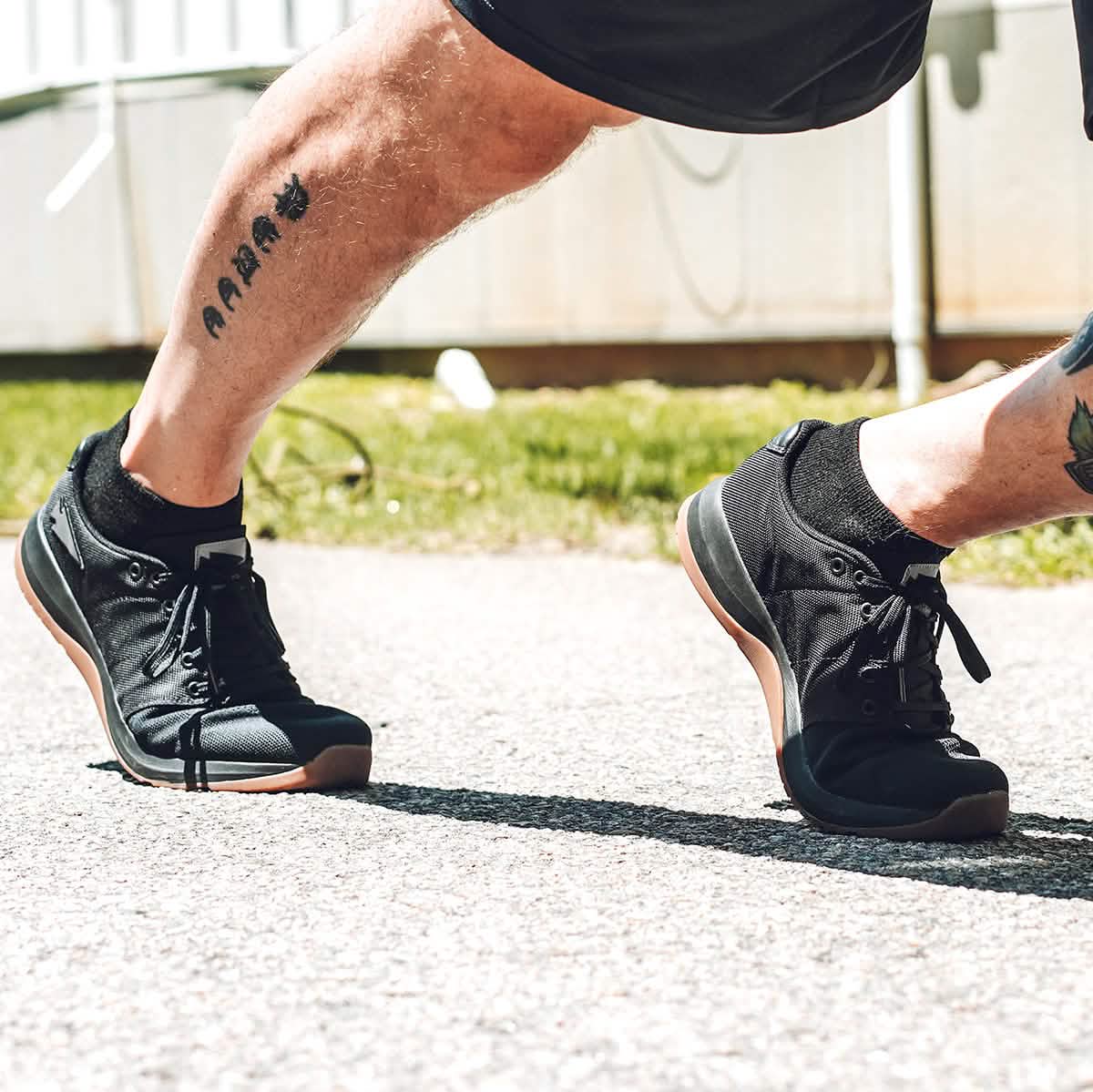 A person wearing GORUCK Men's Ballistic Trainers - HULK and shorts jogs on a sunlit path. The tattooed leg is visible, and the scene suggests an active, outdoor atmosphere.