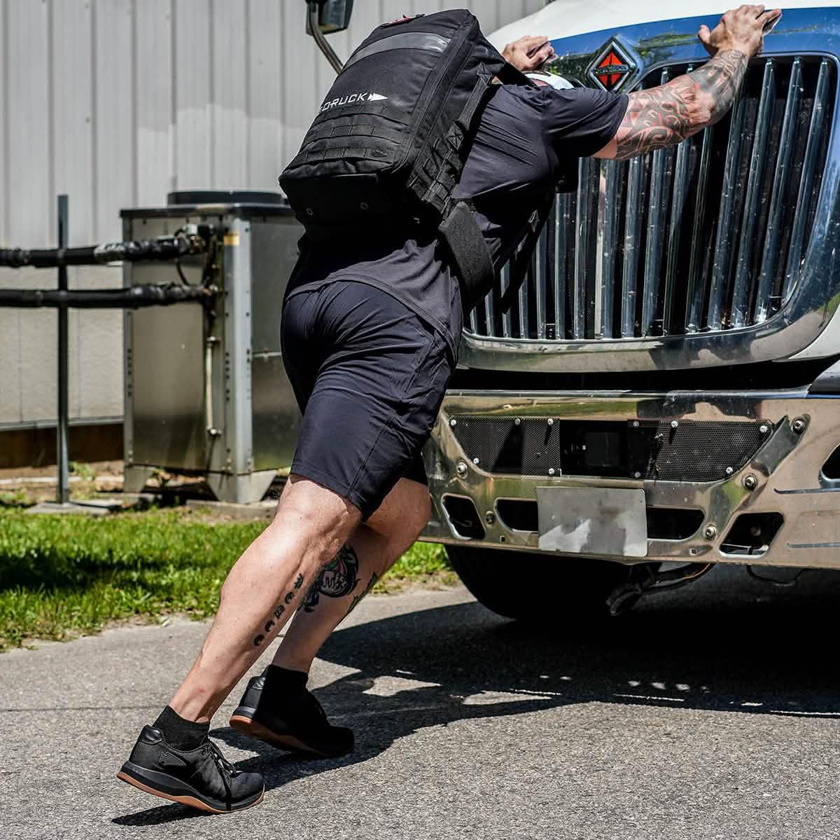 Man wearing a black GORUCK backpack pushing a large truck outdoors, showcasing rugged rucking gear