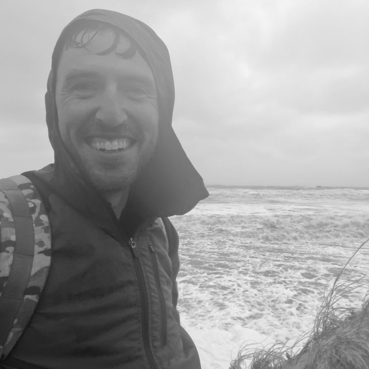 Smiling man in an Indestructible Challenge Windbreaker stands by a rough, windy beach on an overcast day.