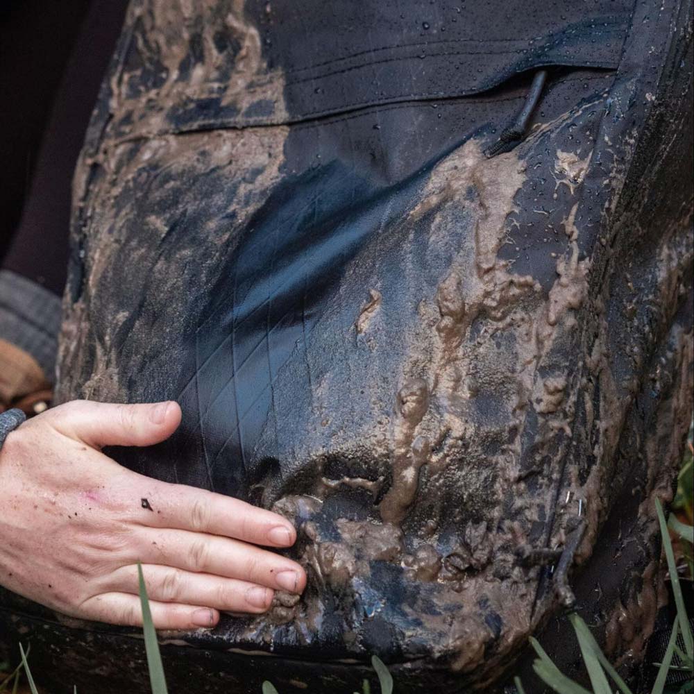A person's muddy hand touching a mud-covered black backpack outdoors.