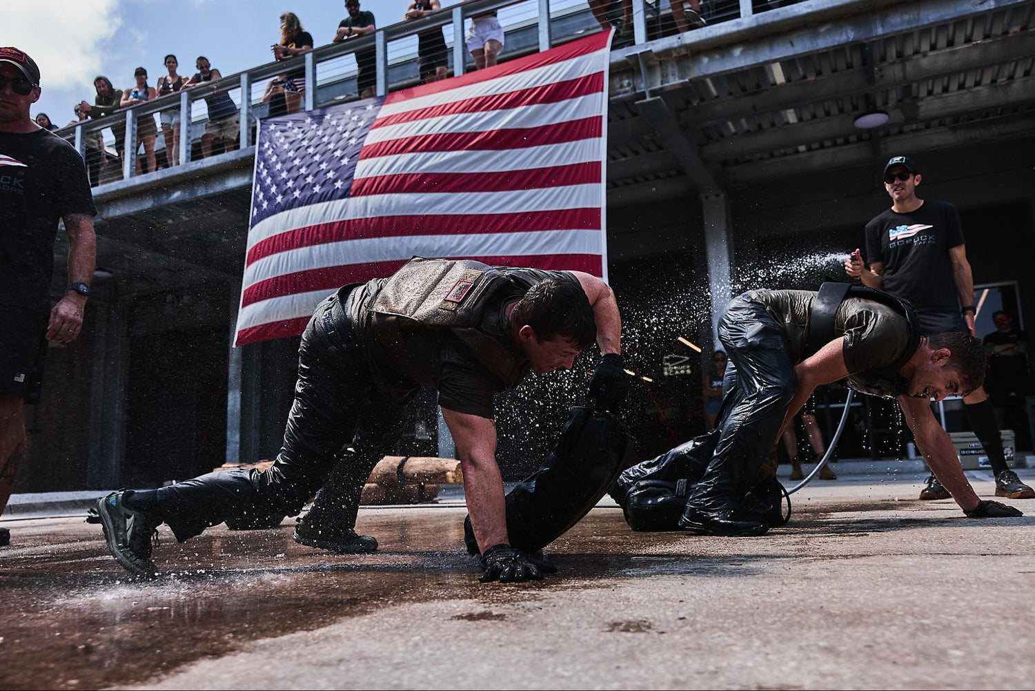 Two people in wet clothes crawl on the ground in front of a large American flag and a crowd.