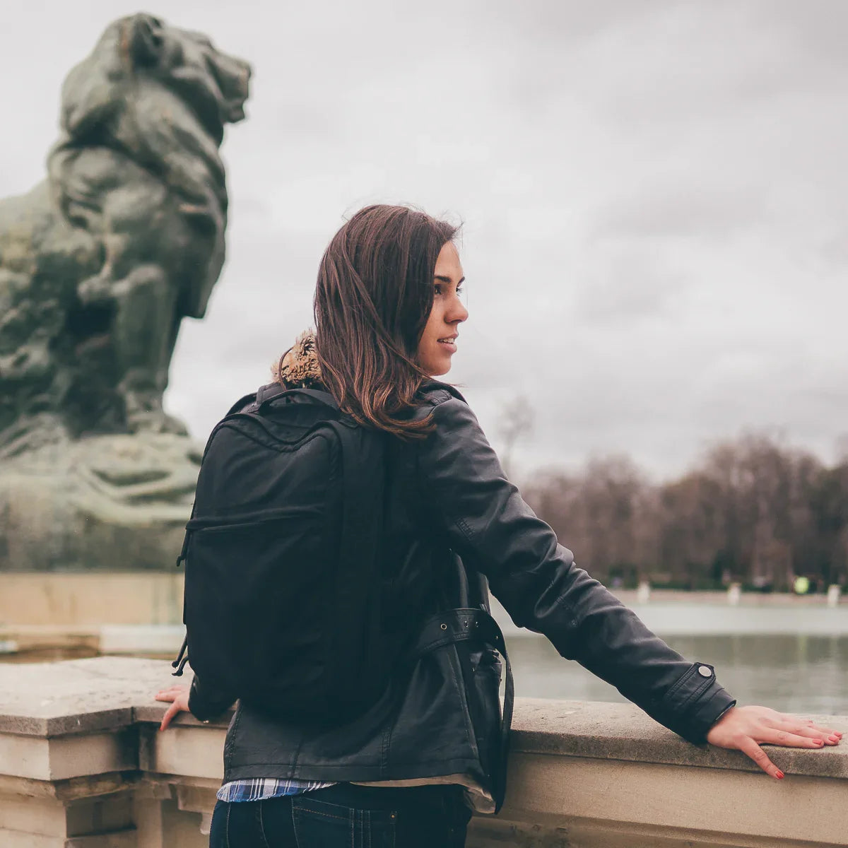 Woman wearing black GORUCK backpack and leather jacket overlooking stone barrier near lion statue by water