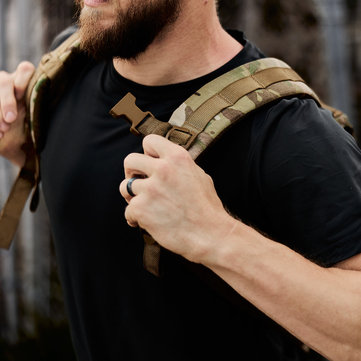Man in black shirt adjusting camo backpack straps, seen from the neck down, outdoors.