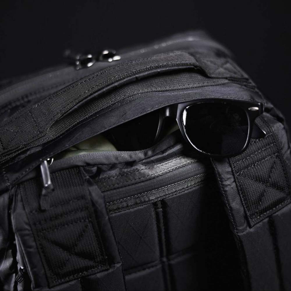 Black sunglasses partially tucked into a zippered pocket of a black backpack against a dark background.
