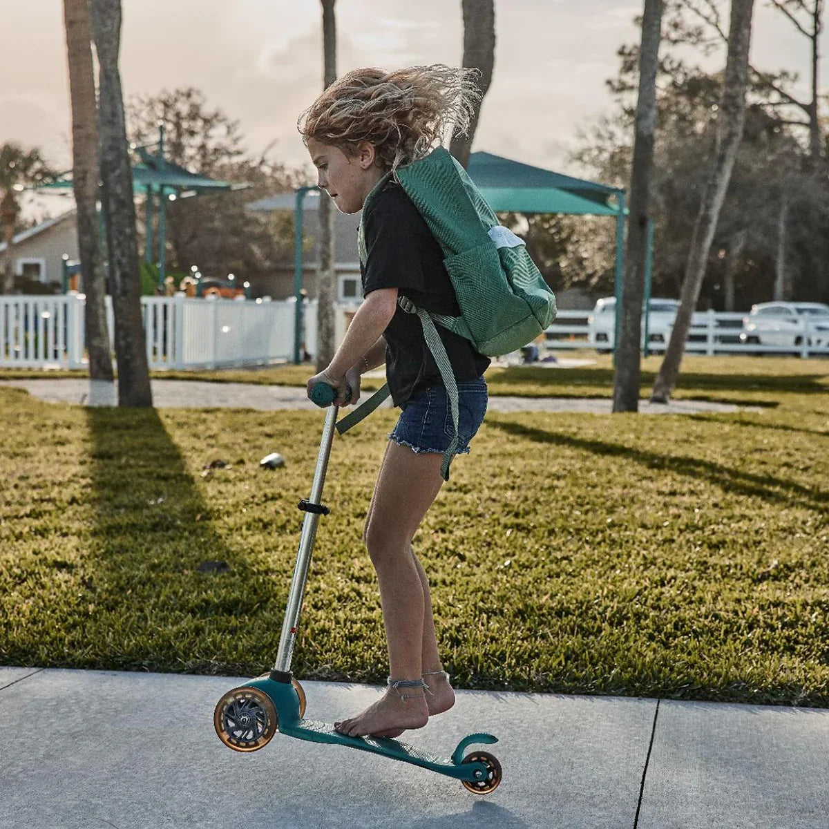 Child with green backpack riding teal scooter barefoot on sidewalk in sunny park