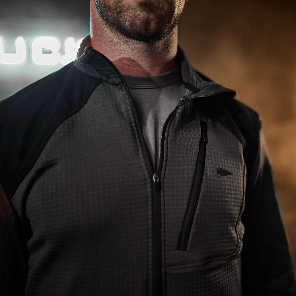 A person wearing a GORUCK Men’s Half Zip Polartec Grid Fleece jacket and a gray shirt is seen in low light. The focus is on the jacket's texture and the person's upper torso, with blurred text in the background.