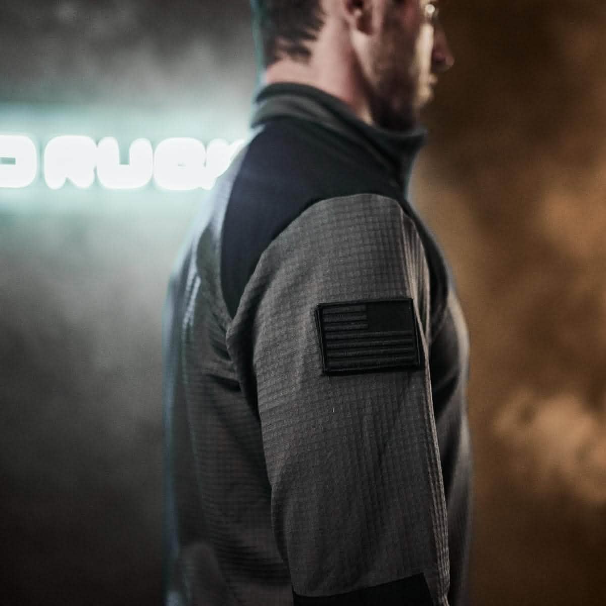 A man wearing the GORUCK Men’s Half Zip - Polartec Grid Fleece stands sideways in a dimly lit room, a patch embellishing his sleeve as a blurred sign glows softly in the background.
