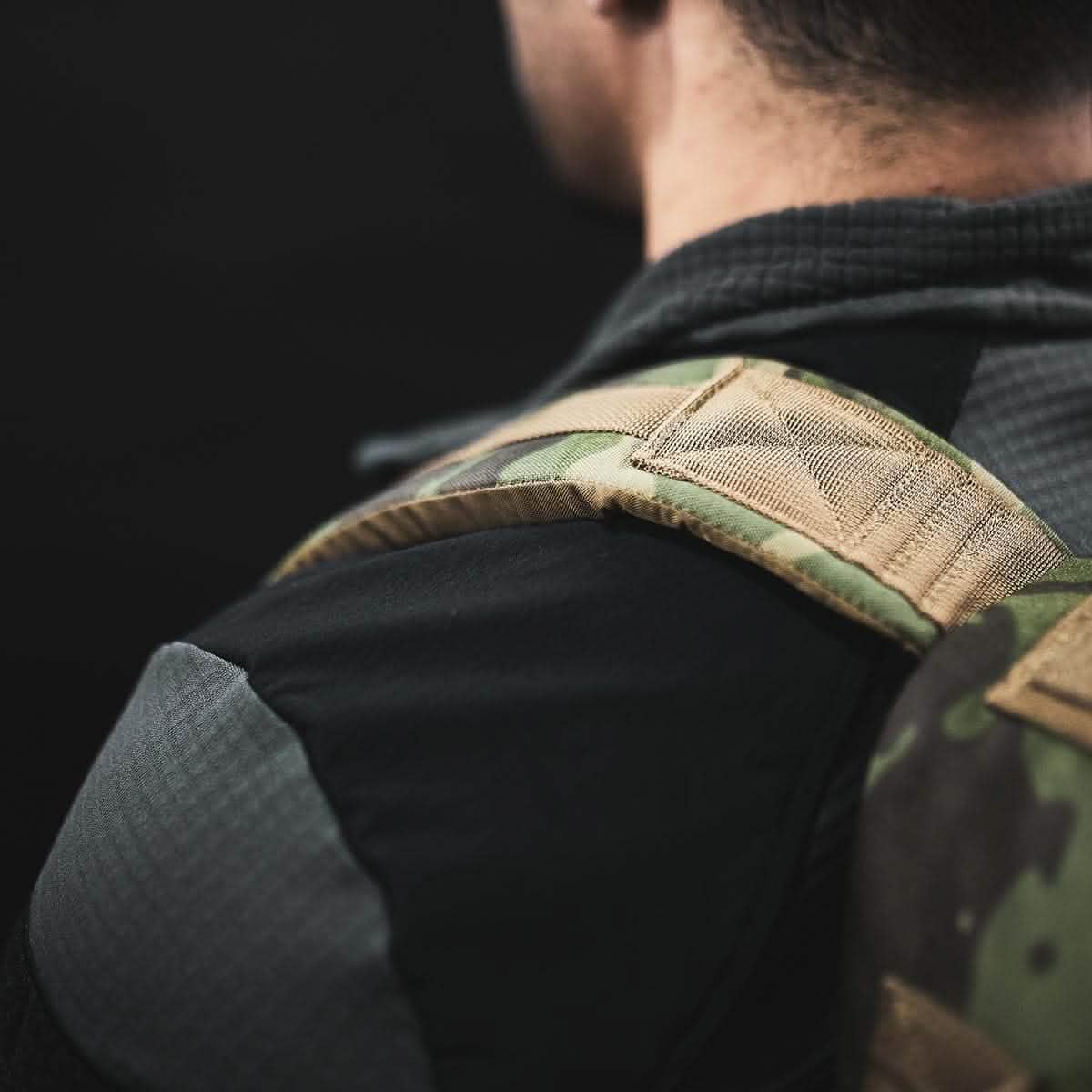 Close-up of a person wearing a camouflage-patterned backpack strap crafted with ToughDry Fabric, paired with the GORUCK Men’s Half Zip - Polartec Grid Fleece in dark clothing. The individual is facing away from the camera against a black background, revealing only the shoulder and back of the head.