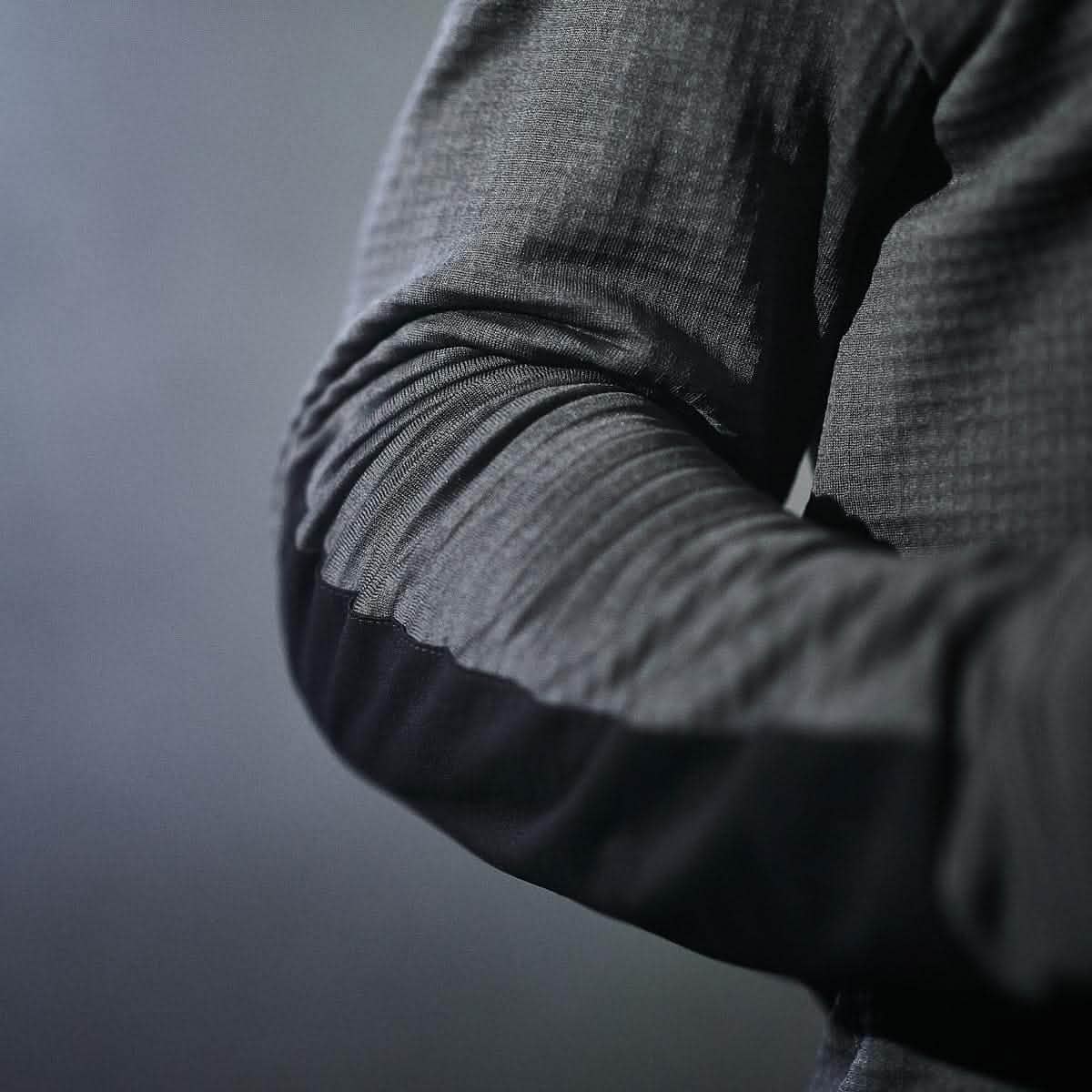 In the foreground, a person is seen wearing the GORUCK Men's Half Zip - Polartec Grid Fleece, characterized by its textured, dark fabric and long sleeves. One arm is crossed over their chest, accentuating the design. The moody, atmospheric background plays off the fleece-lined fabric nicely, offering a subtle contrast against the soft, blurred gray scenery.