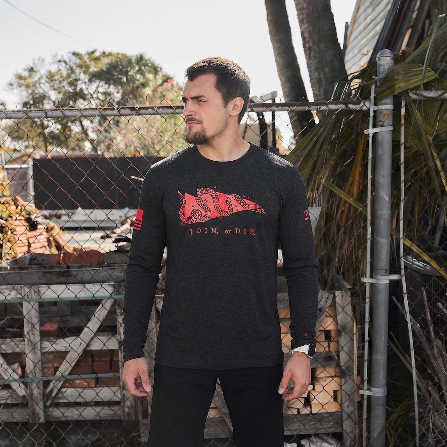 Man standing outside by a fence, wearing the Men's Join or Die Long Sleeve - Tri-BlendX, a classic symbol of colonial unity, looking to the side.