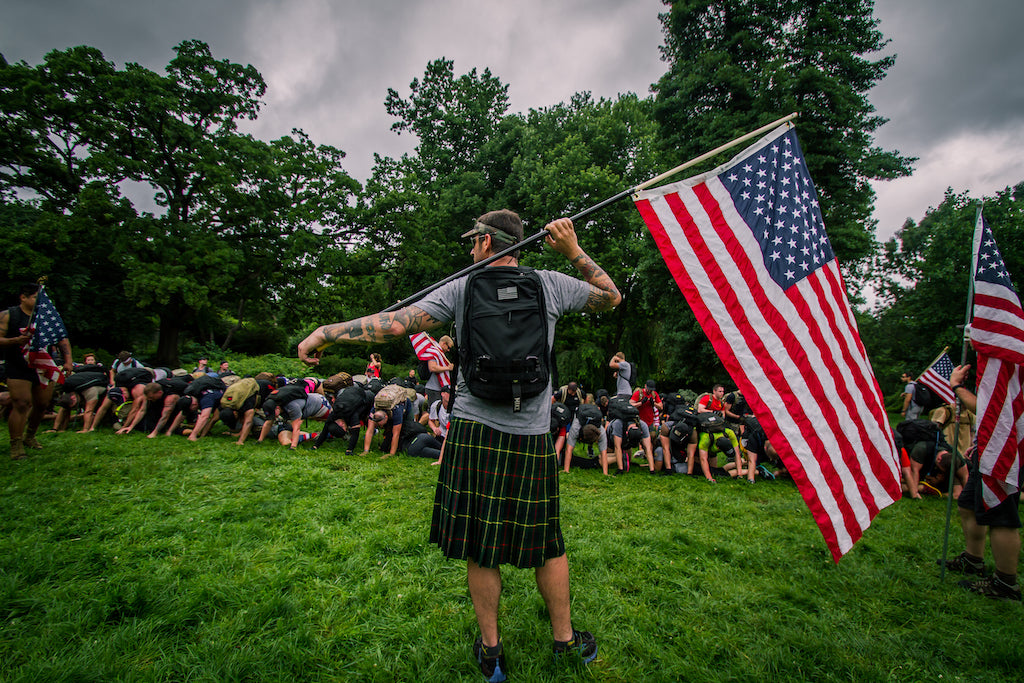 A person in a kilt holds an American flag while a group bends down on a grassy field under cloudy skies.