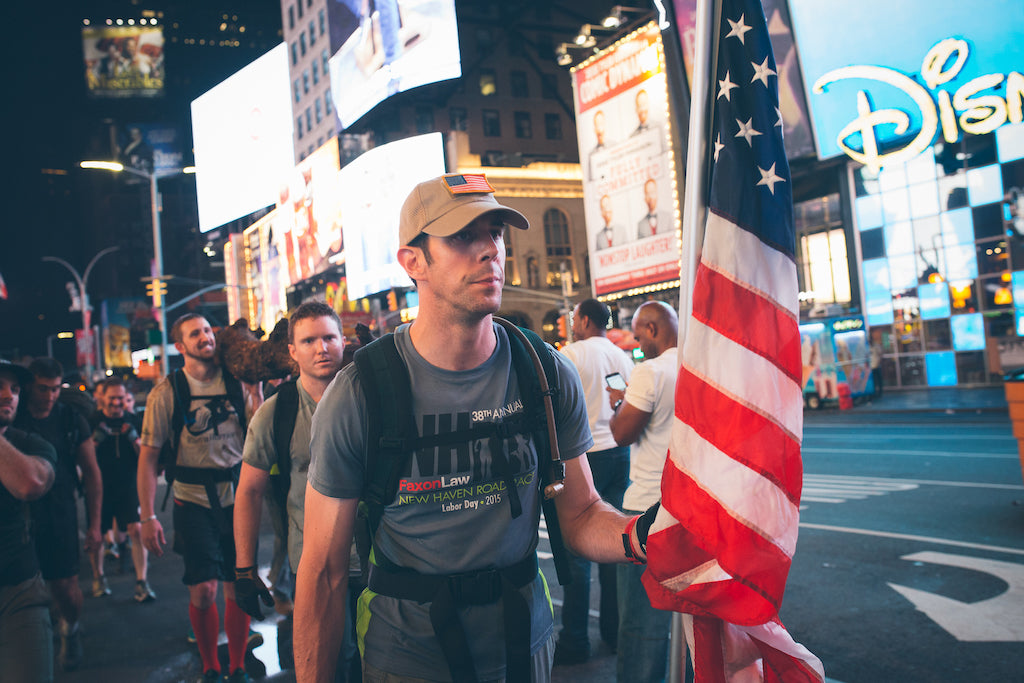 A man carrying a U.S. flag walks with a group in a brightly lit city street at night.