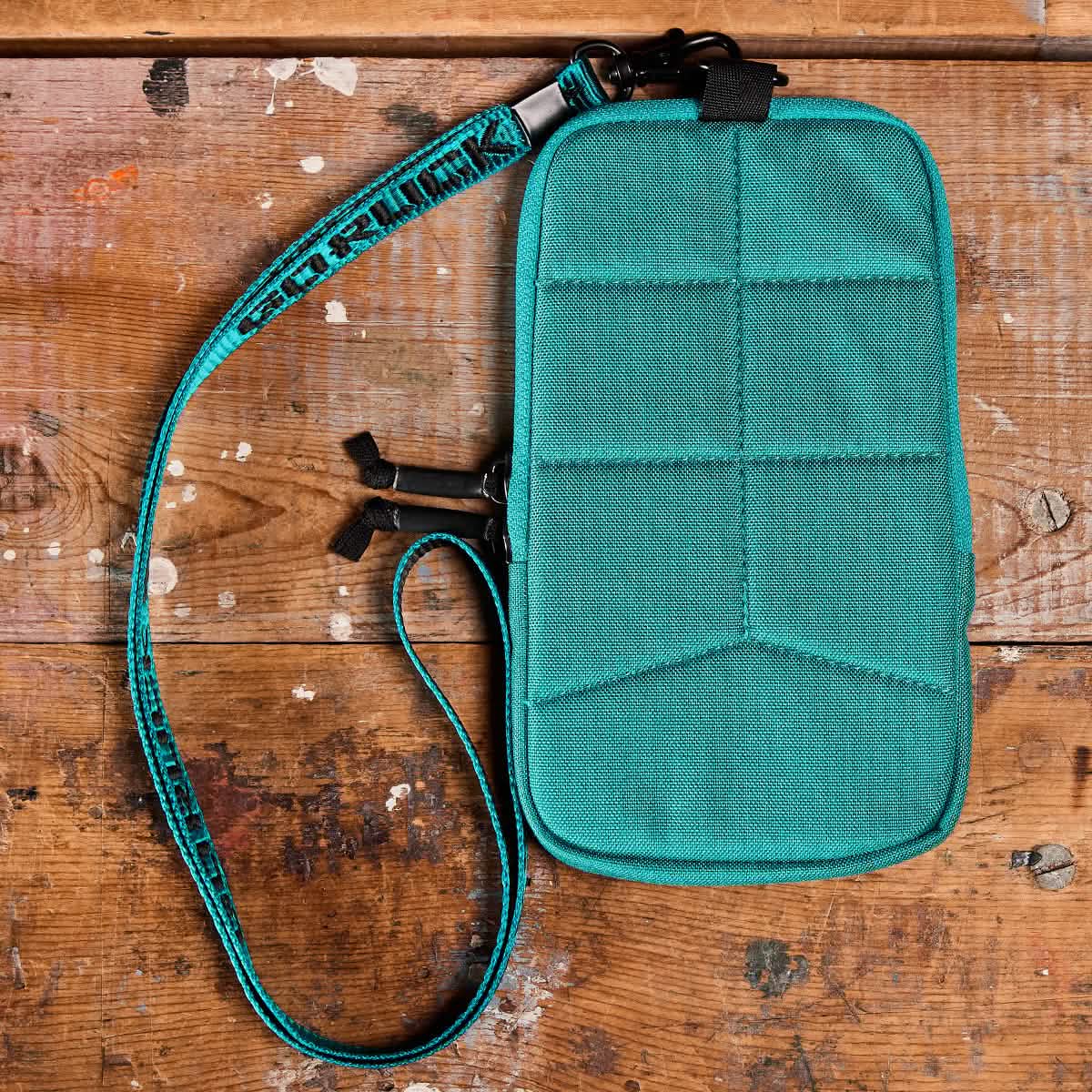 A GORUCK Phone Pouch, made from teal CORDURA fabric in a rectangular shape, is displayed on a wooden surface. It has a zipper with two pulls and showcases a stitch pattern. Accompanied by a matching adjustable strap with black clips, the phone pouch contrasts beautifully with the wood, which is marked by paint stains and scratches.