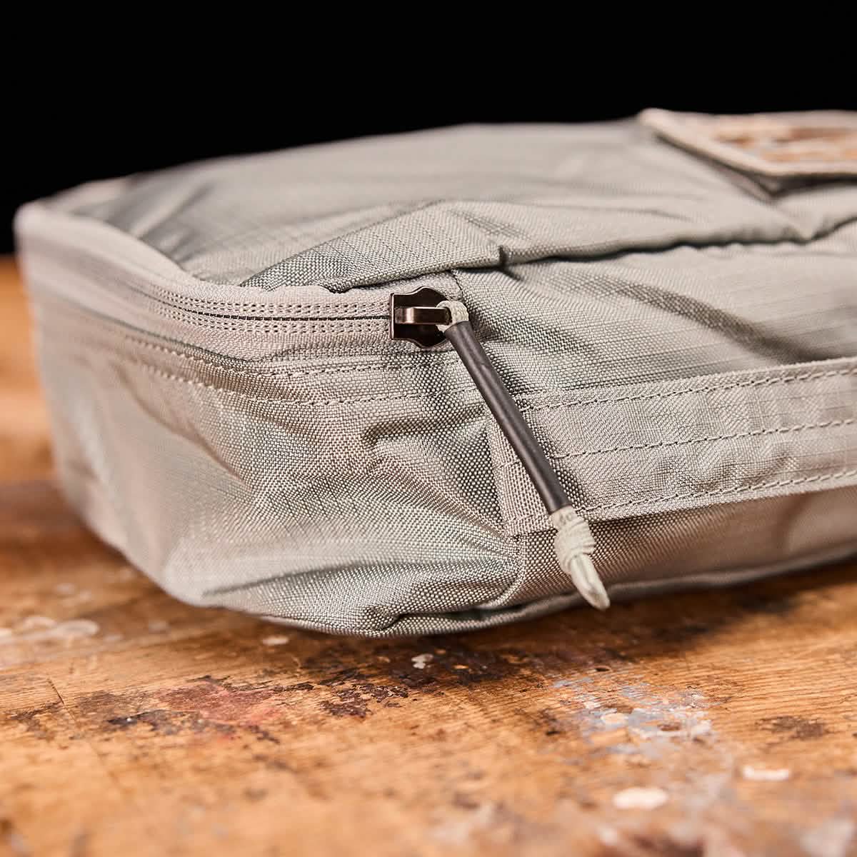 A close-up of a Shadow Pocket by GORUCK, crafted from rugged ROBIC Ripstop Nylon in gray, featuring a zipper resting on a wooden surface. The zipper pull is visible, highlighting the textured material. Ideal for organizing your rucksack, it stands out against the black background.
