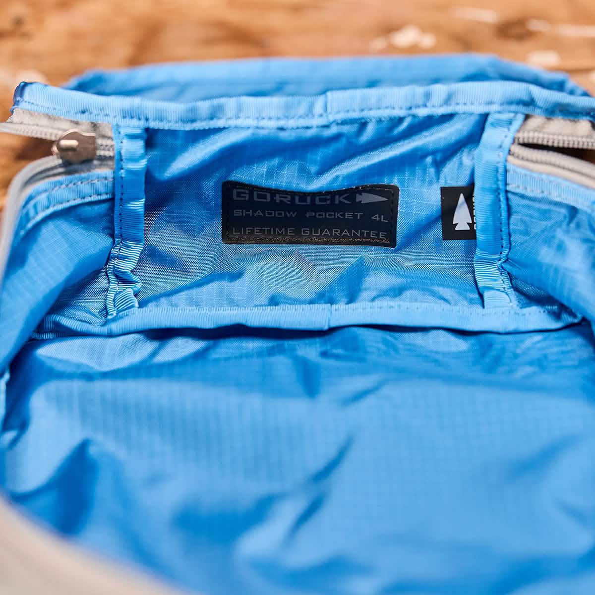 A close-up of a blue GORUCK Shadow Pockets - Ripstop ROBIC® 4L made from durable ROBIC Ripstop Nylon, featuring a visible label that says Lifetime Guarantee. The pocket is open to reveal its roomy interior for optimal rucksack organization, set against a wooden surface background.