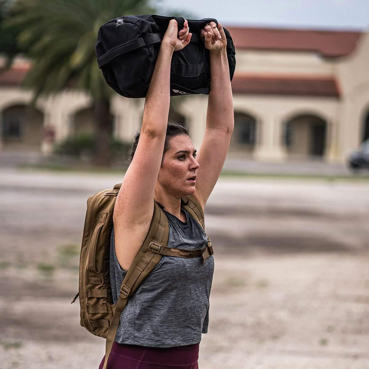 Woman rucking outdoors in GORUCK light grey performance tank, lifting sandbag, with backpack