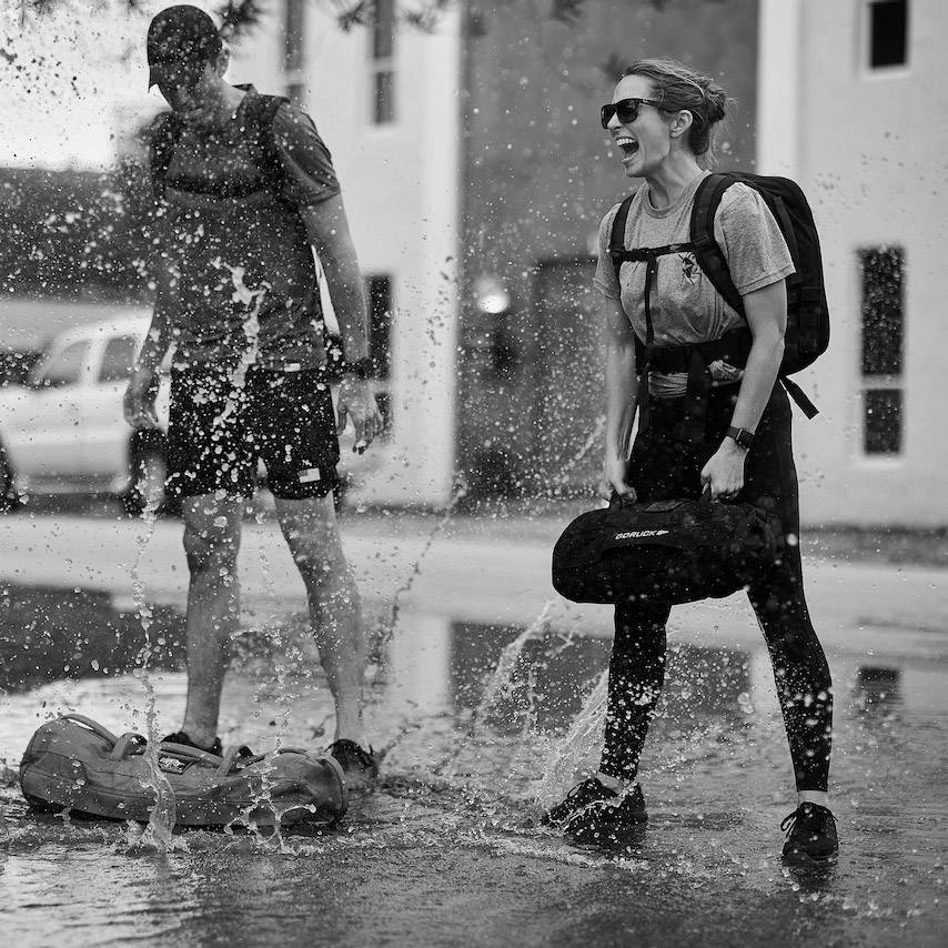 Two people splash in a puddle, laughing, wearing backpacks and casual clothes. Black and white photo.