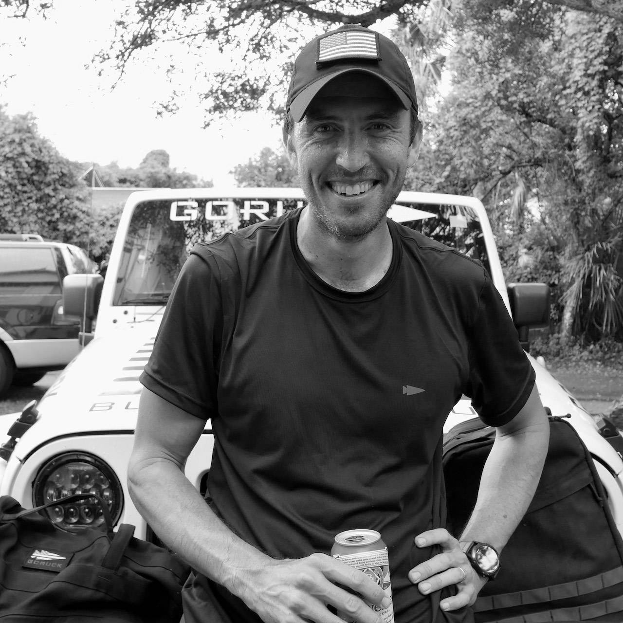 Man in a cap smiling, holding a can, standing in front of a jeep with trees in the background.