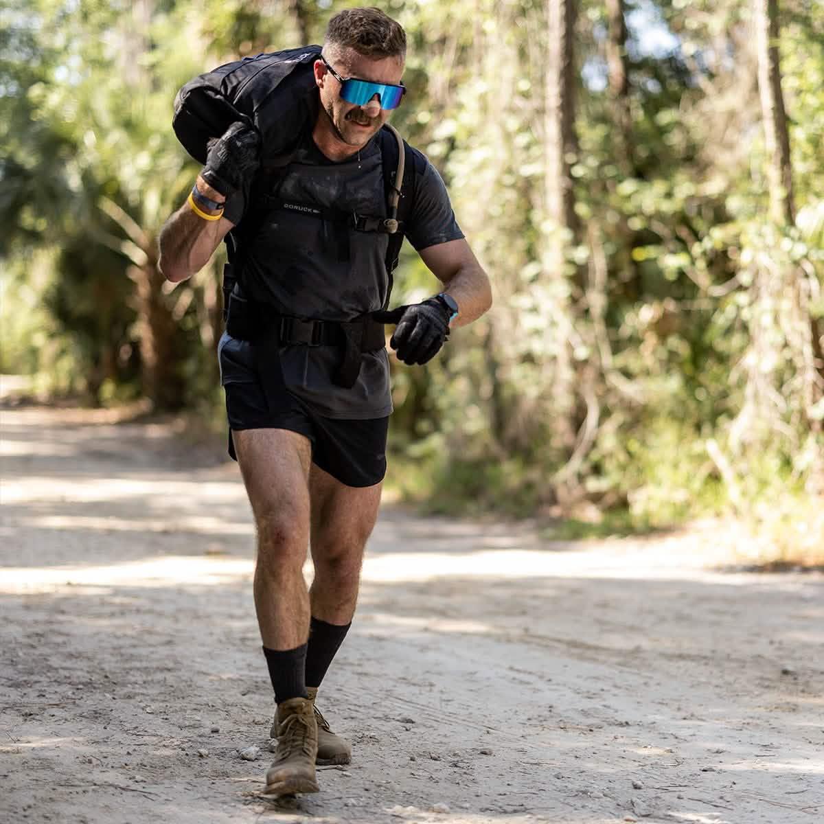 Man wearing sunglasses and GORUCK gear carrying a heavy rucksack on a forest trail