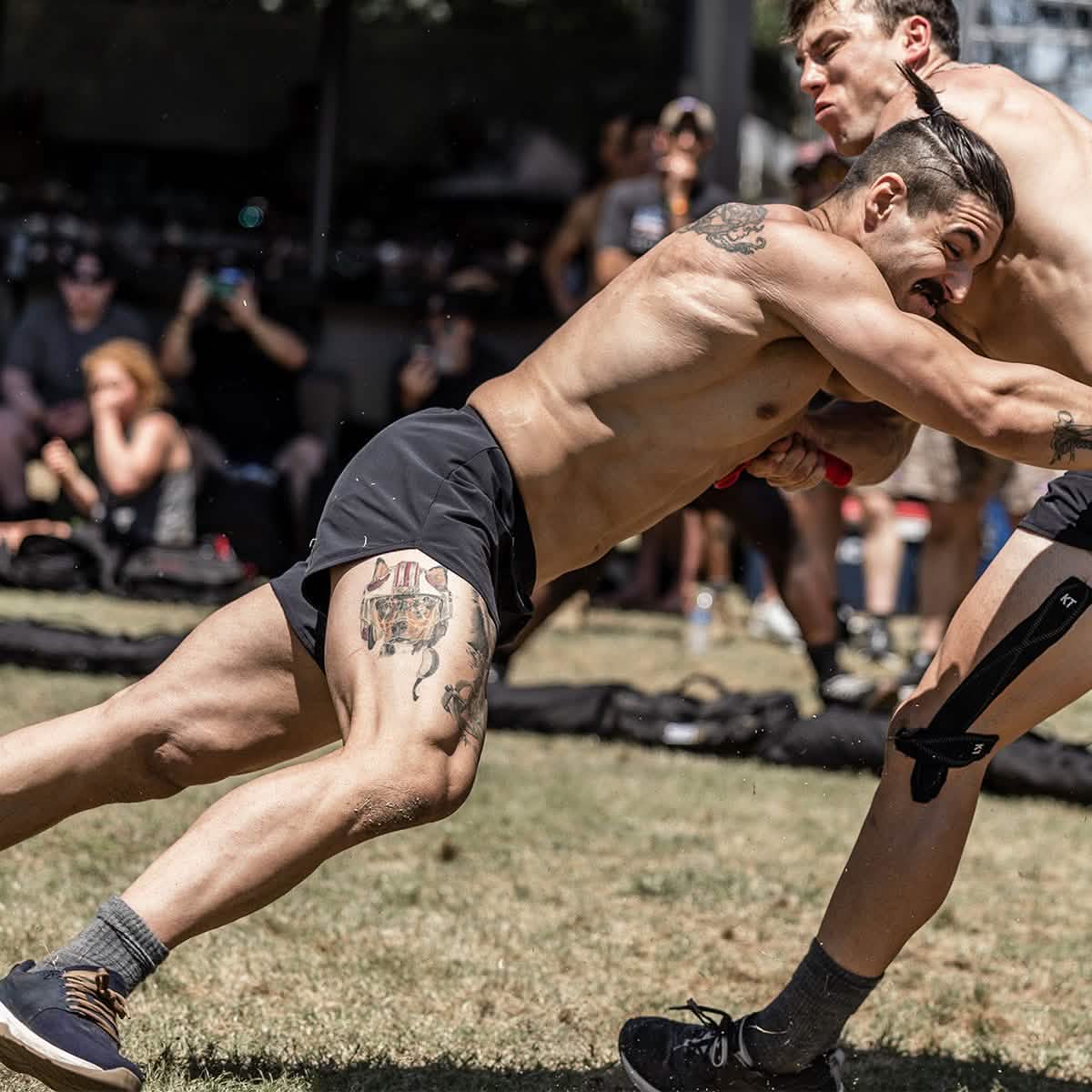 Two men wearing GORUCK's Men’s Ranger Panties - ToughStretch engage in an outdoor wrestling match on the grass. One, adorned with tattoos and wearing a knee brace, highlights the durability of ToughStretch fabric; the other sports short hair. Enthusiastic spectators look on eagerly, capturing moments with their cameras. The scene is lively and spirited, exemplifying a rugged essence backed by Scars Lifetime Guarantee confidence.