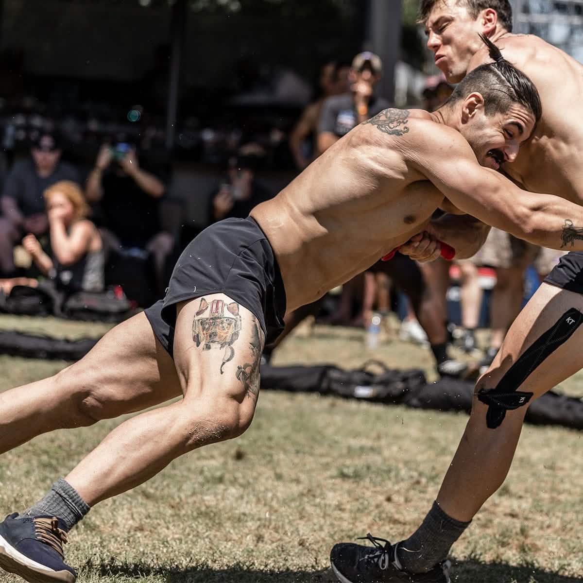 Two shirtless men wrestling outdoors on grass with spectators blurred in the background