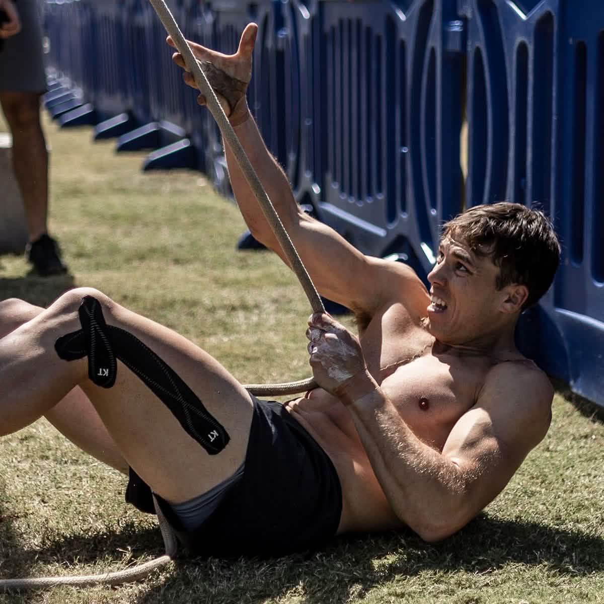 Shirtless man with athletic build pulling on rope while lying on grass near blue barriers during outdoor workout