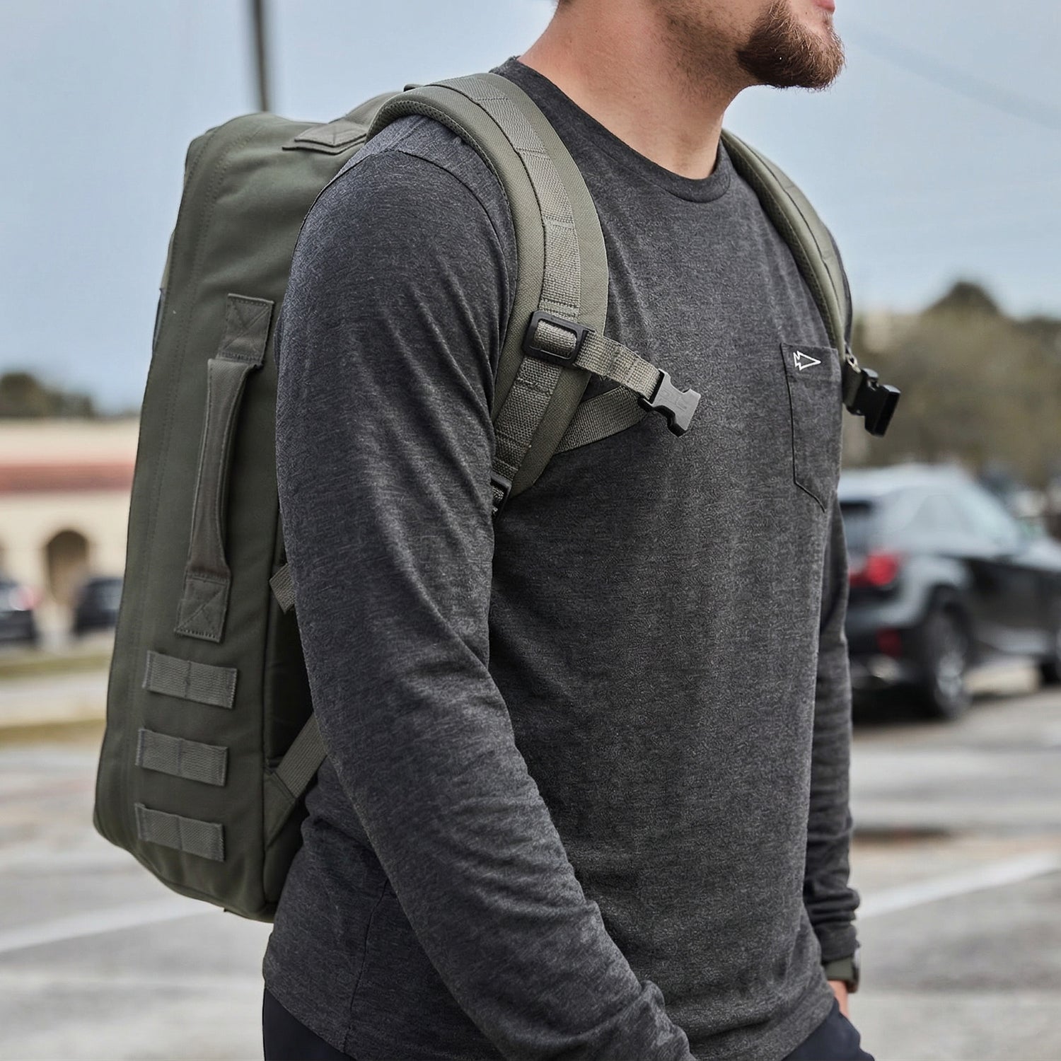 Man wearing a Men's Long Sleeve Pocket Tee - Tri-BlendX in gray, carrying a large green backpack outdoors in a parking lot.