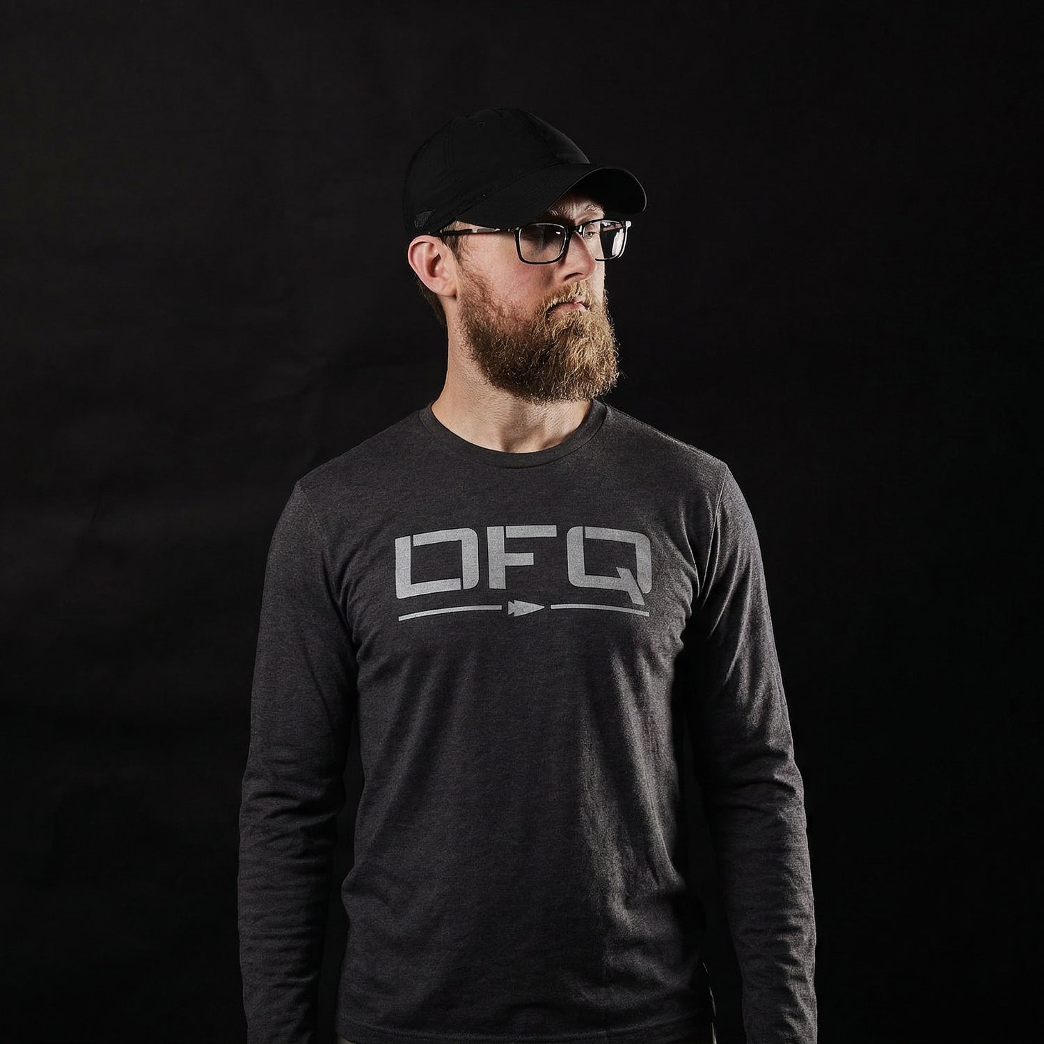 Bearded man with glasses wears a black cap and a dark Men's DFQ Long Sleeve - Tri-BlendX shirt, standing against a black background.