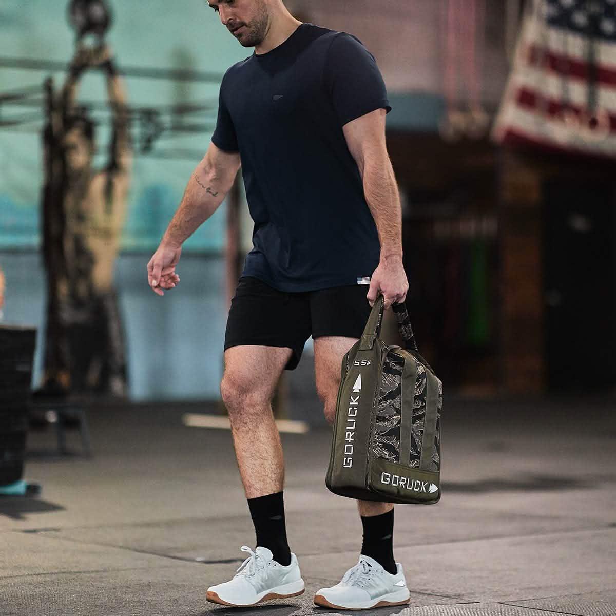 Athletic man in gym carrying GORUCK Lunar Rock bag with camouflage pattern and 55 lb weight label