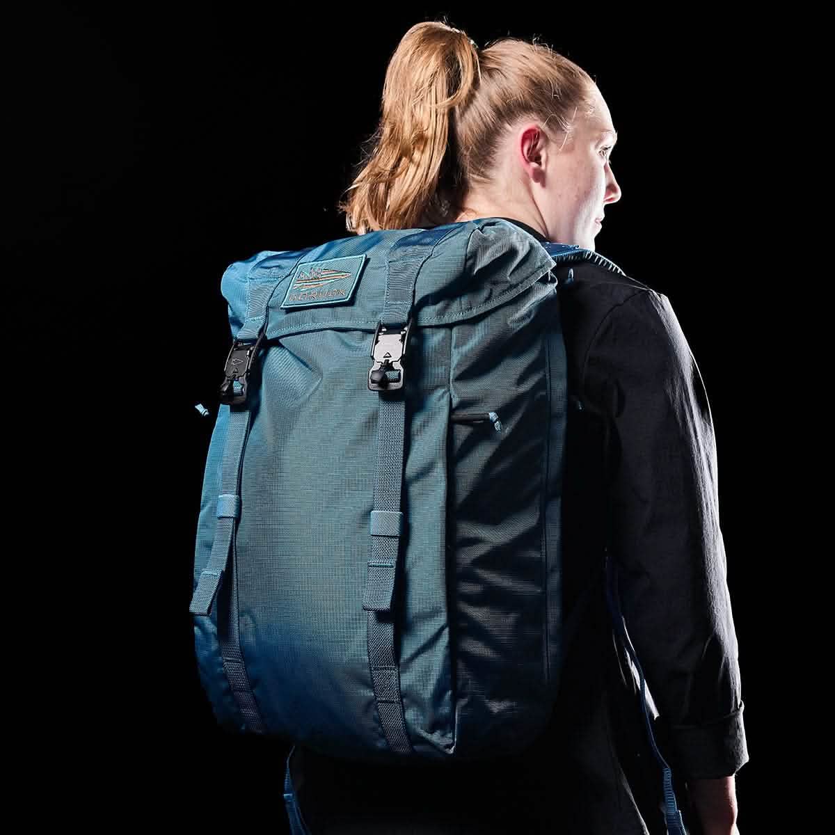 A person with long hair is wearing a GORUCK M23 backpack in Tidal Blue + Speed Tan, featuring a military-style with multiple buckles and textured Ripstop ROBIC® fabric adorned with a logo patch. Wearing a black outfit, the background complements the 21L rucksack.