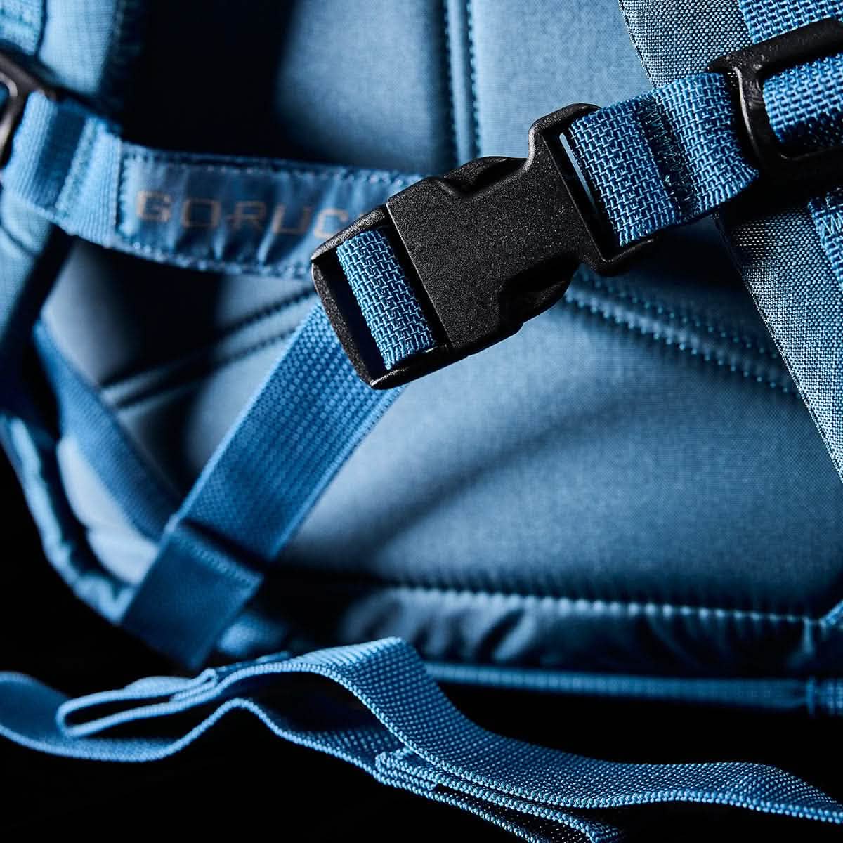 Close-up of the GORUCK M23 harness, crafted from Tidal Blue Ripstop ROBIC® fabric with a Speed Tan plastic buckle, showcasing detailed stitching reminiscent of an M1945 field pack. The harness is set against a dark background, highlighting the vibrant blue color and the contrast between the fabric and the buckle.