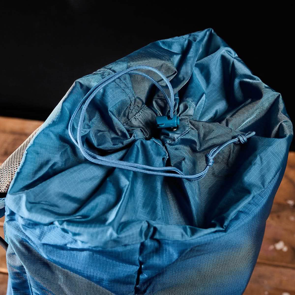 A close-up of the GORUCK M23 military backpack in Tidal Blue and Speed Tan, with its top drawstring closure open, revealing the fabric and cords. The 21L rucksack rests on a wooden surface against a dark background.