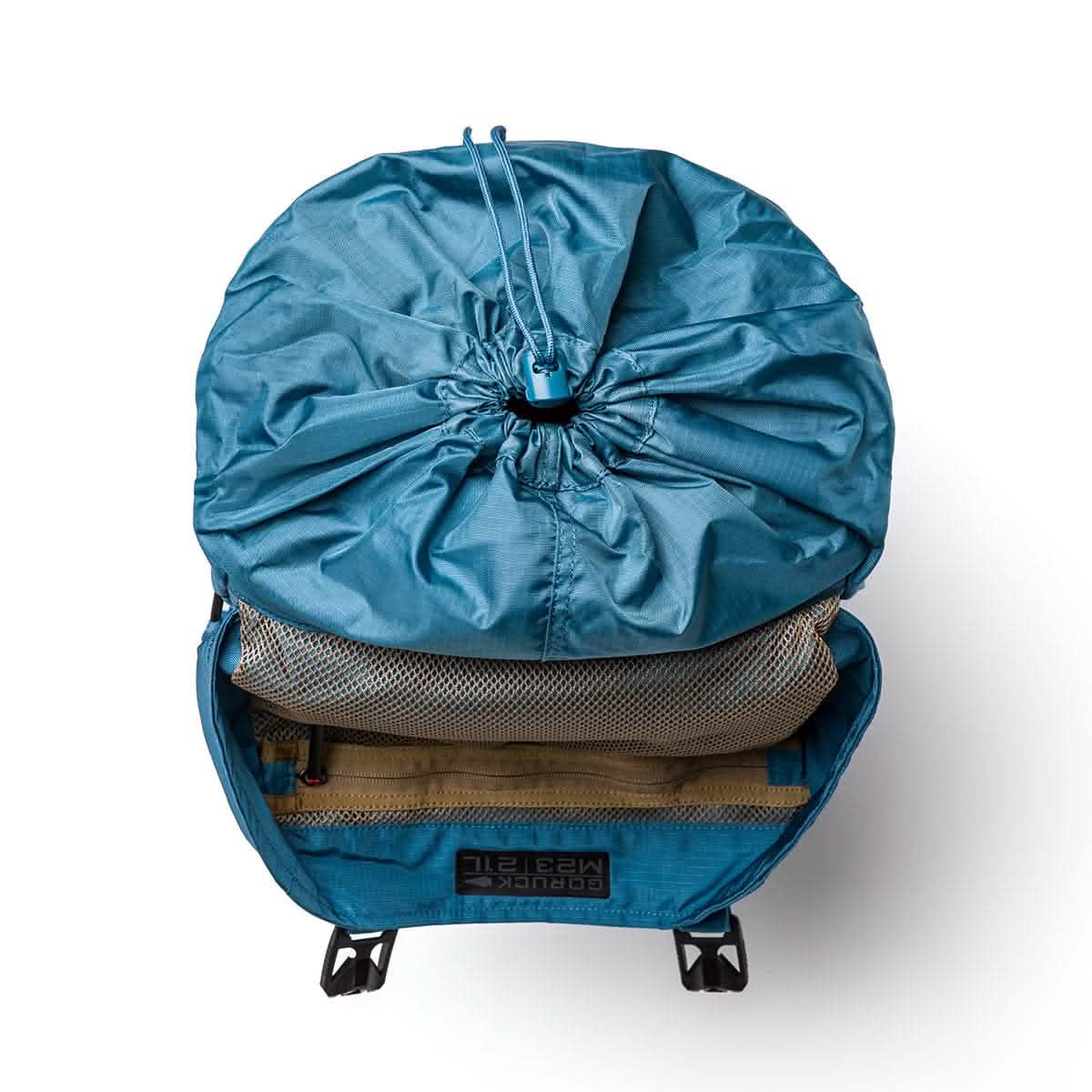 A tidal blue rucksack with its lid open reveals a drawstring closure and mesh pocket. The interior features a speed tan compartment for organization. The brand GORUCK is visible at the bottom, adding a rugged touch reminiscent of a vintage M1945 field pack.