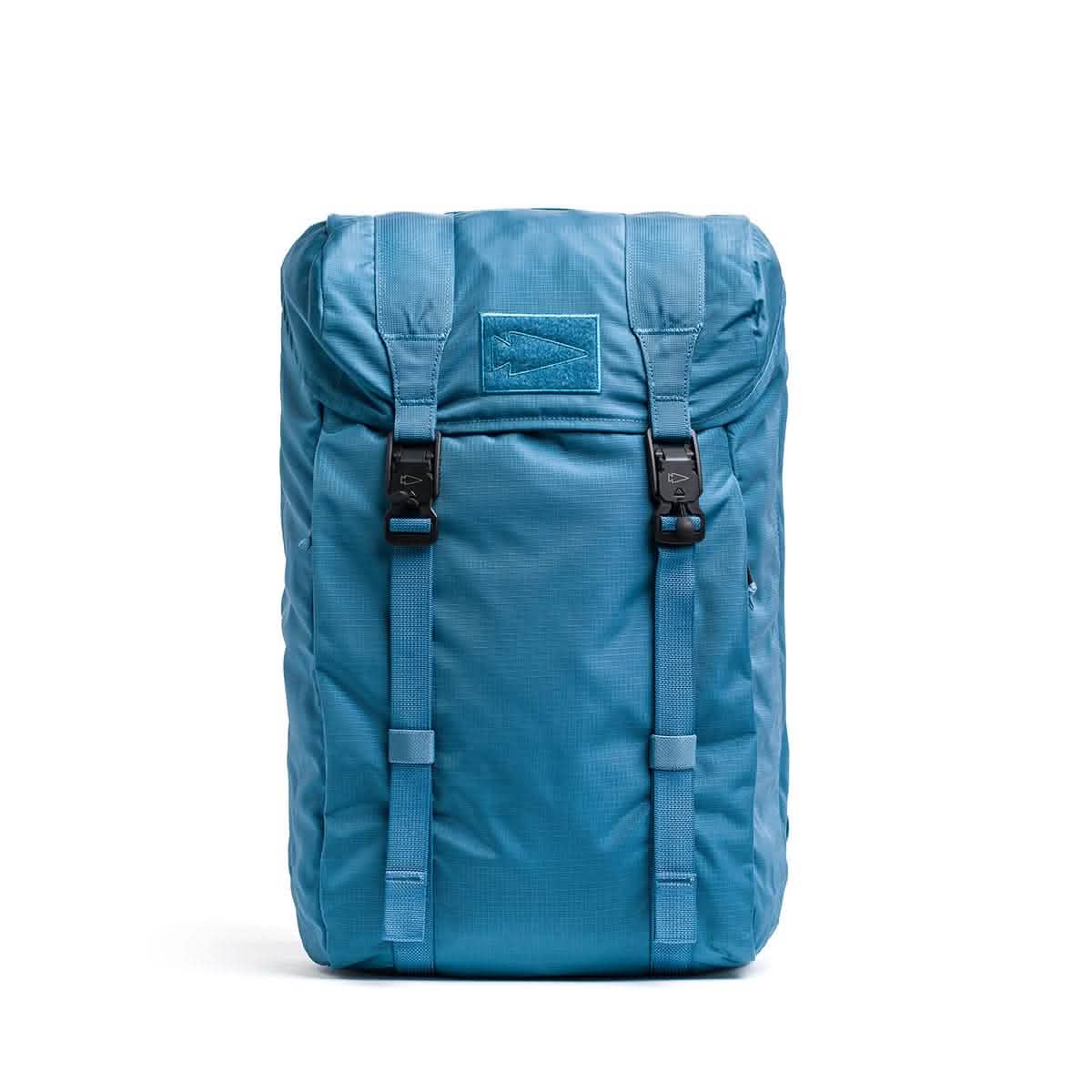 The GORUCK M23 - Ripstop ROBIC® in Tidal Blue and Speed Tan is a 21-liter rucksack with a top flap secured by two black buckles. It features a subtle stitched logo on the front and is made from durable fabric reminiscent of military backpacks, making it ideal for outdoor activities or travel.