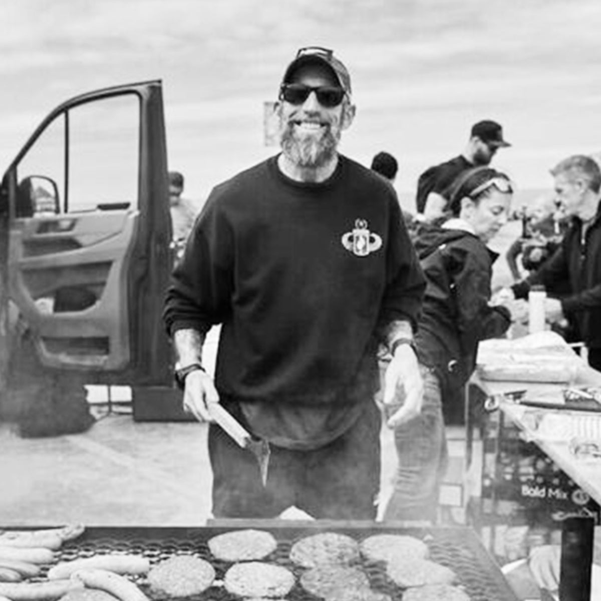 A bearded man smiles while grilling burgers and sausages at an outdoor gathering with people in the background.