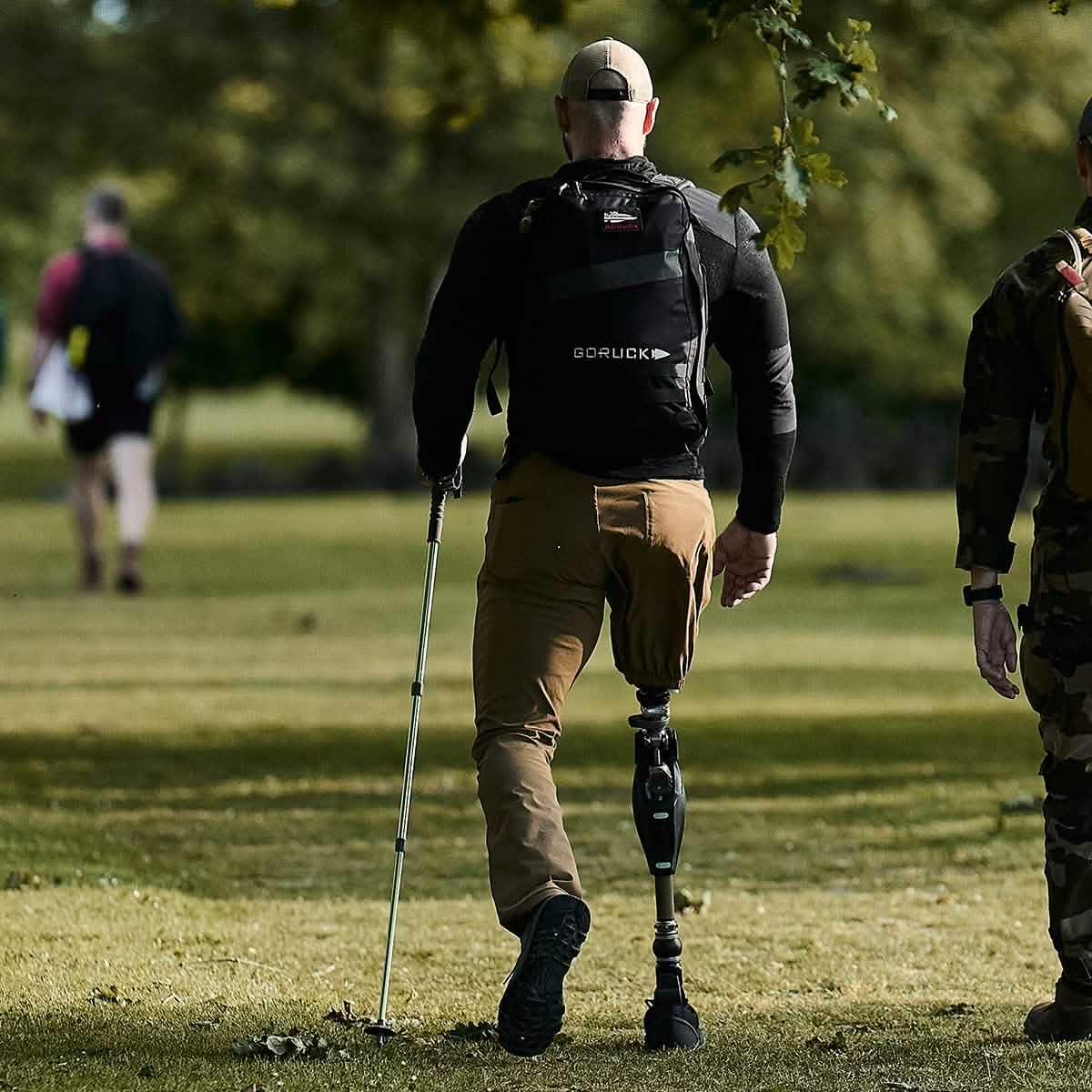 Man with prosthetic leg carrying a GORUCK backpack walking outdoors on grass with others