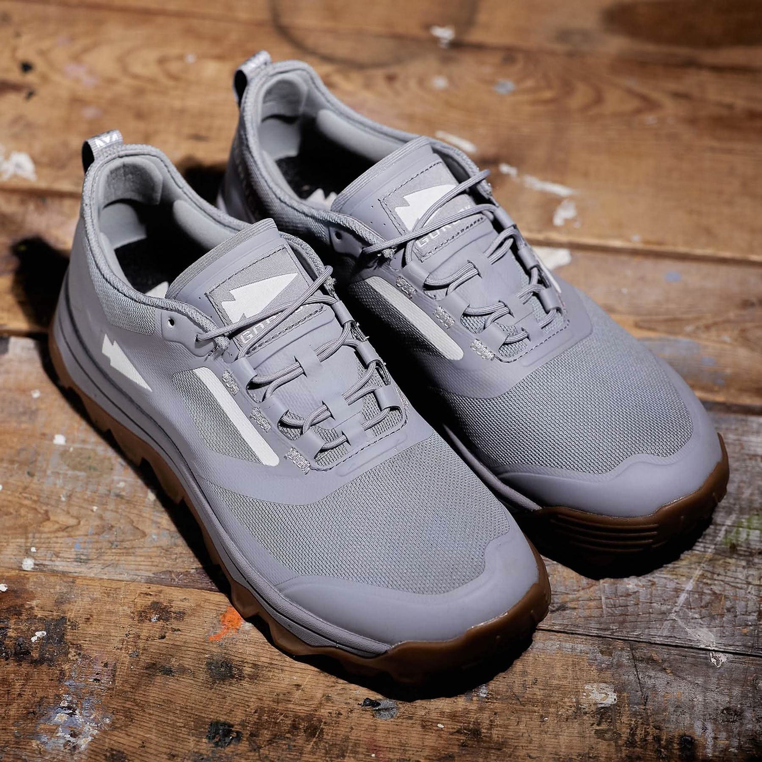 A pair of Mackall - Chiseled Stone + Gum by GORUCK rests on a wooden surface. These sleek rucking shoes feature a mesh fabric, lace-up closure, and a triple compound outsole. The lighting beautifully highlights their texture and details.