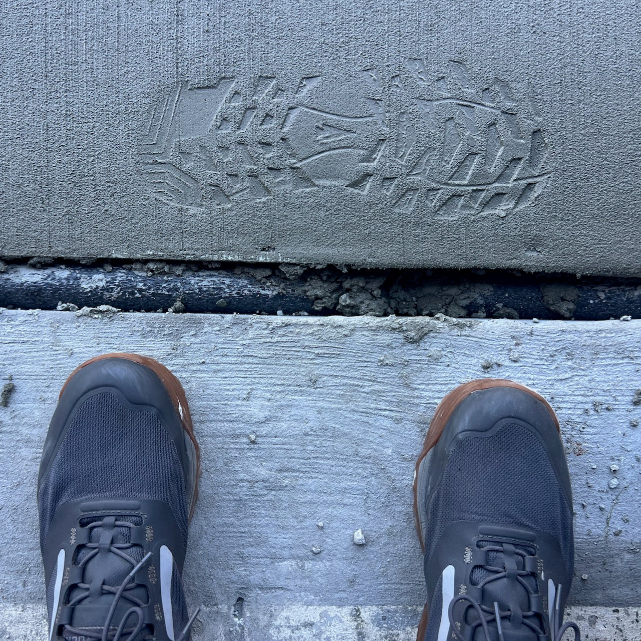 A shoe print is visible in wet cement, with a pair of Mackall - Forged Iron + Chiseled Stone + Gum trail shoes standing nearby on dry concrete.