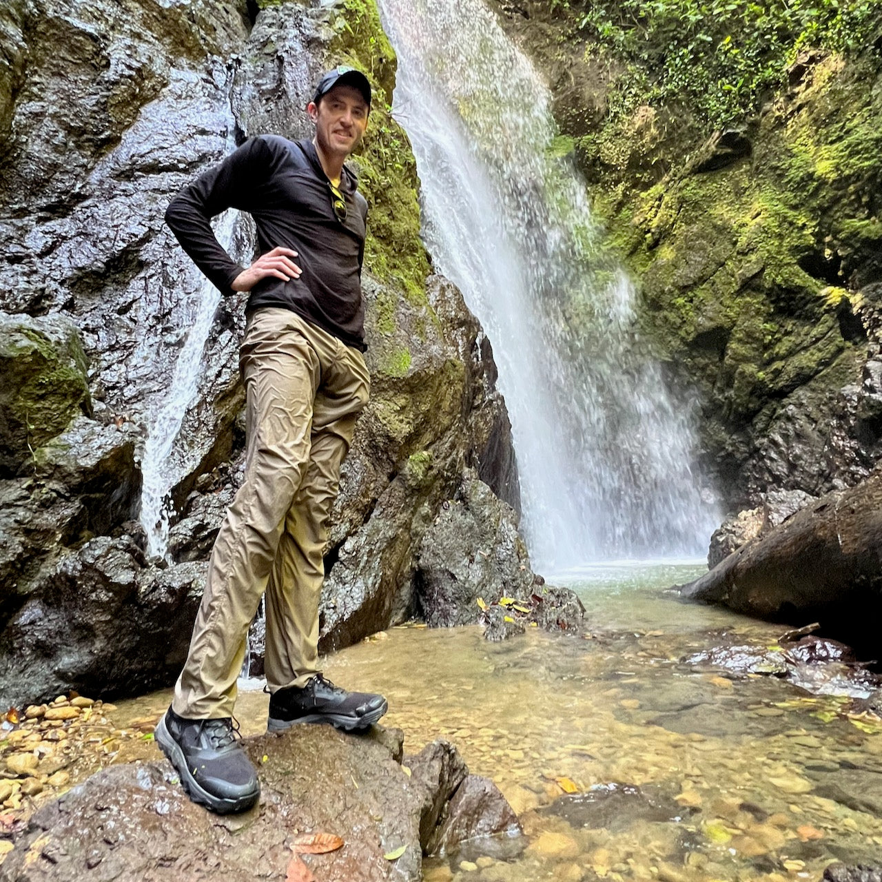 Smiling at the camera, a person stands on rocks beside a waterfall in a lush green forest, wearing Mackall - Black + Forged Iron all-terrain trail shoes.