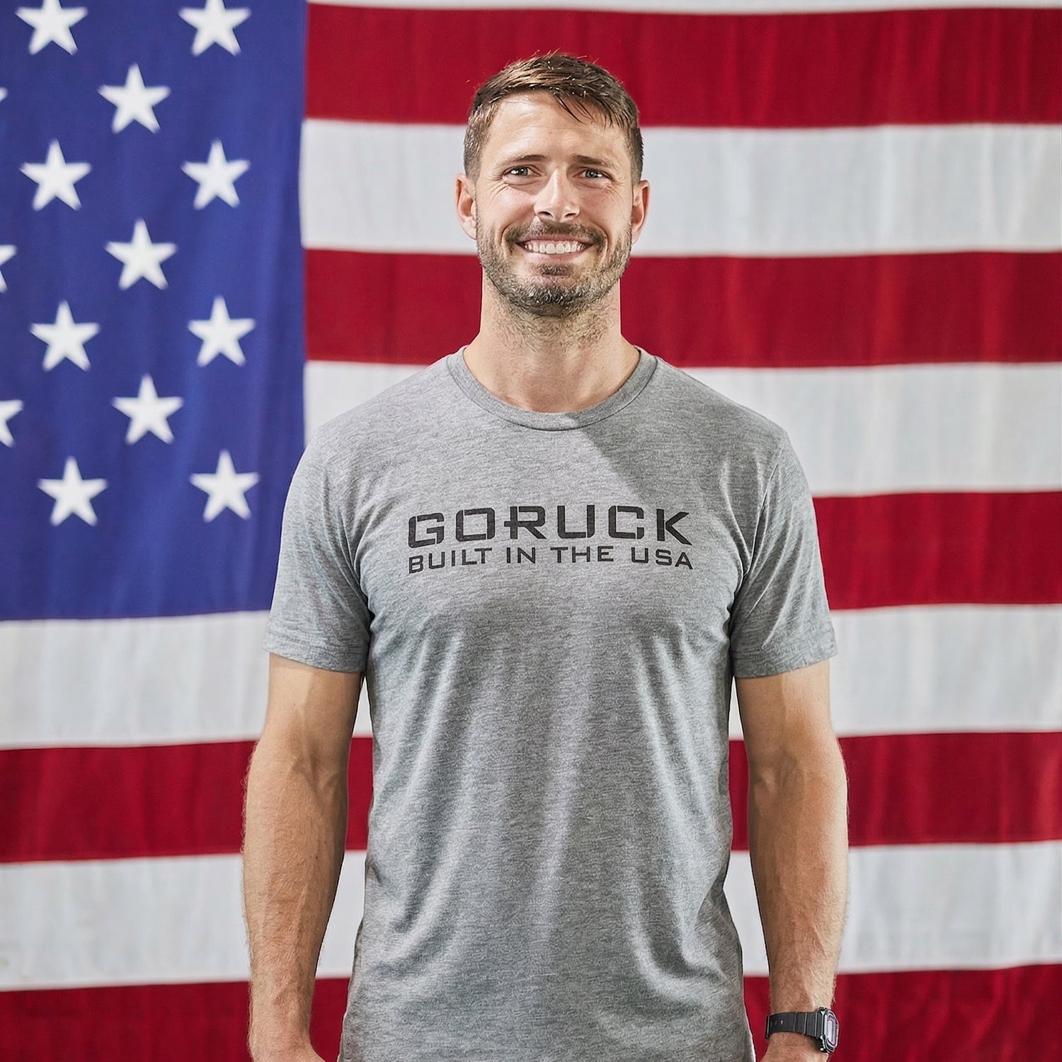A smiling man wears the GORUCK Built in the USA Tee - Tri-BlendX, featuring a premium polyester-cotton-rayon blend, as he stands before a large American flag—symbolizing durable American-made gear.