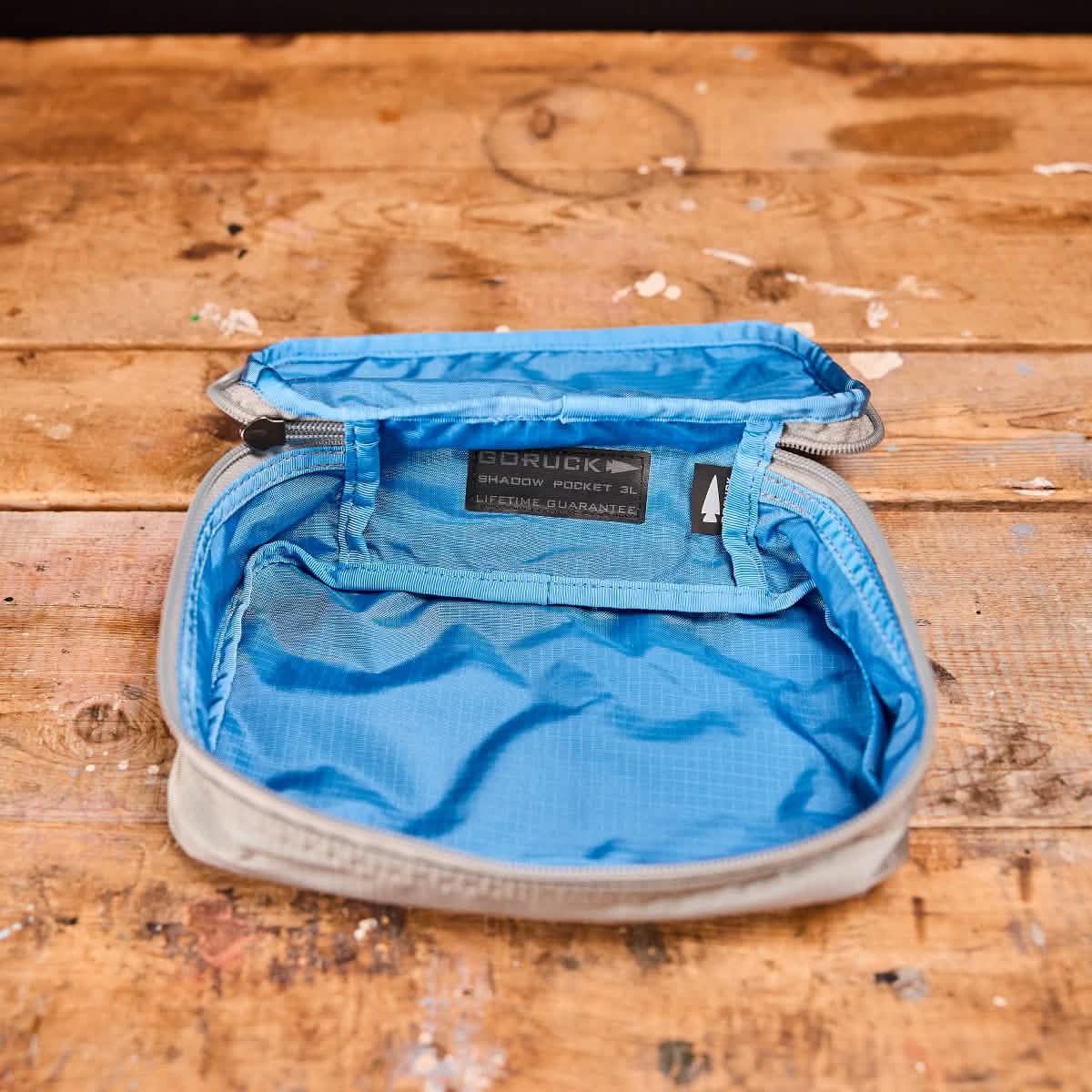 A Shadow Pocket from GORUCK, featuring an open design in light grey and blue, crafted from 420D Ripstop ROBIC® Nylon, sits on a wooden surface. Perfect for organizing within your rucksack, its blue interior includes a mesh pocket. The lid displays a black tag with text, highlighting the utility of the Shadow Pockets collection.