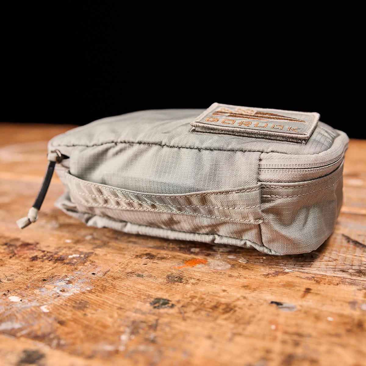 A compact olive-green Shadow Pocket from GORUCK sits on a wooden surface, made with durable Ripstop ROBIC® 420D Nylon. It boasts a patch with text on top and an outer compartment featuring a strapped zipper pull. The slightly rugged texture is ideal for organizing rucksacks and outdoor adventures.