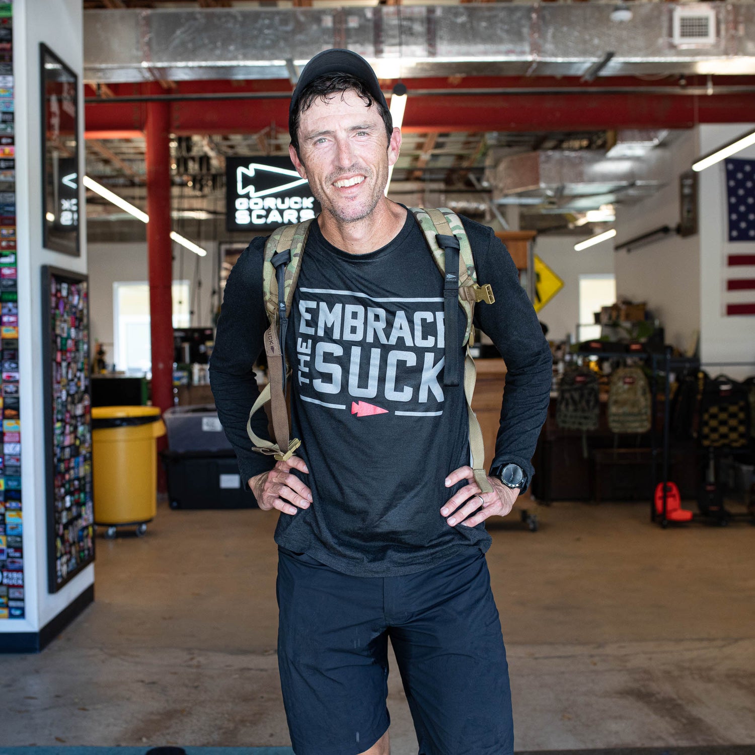 Man in athletic gear with a backpack stands smiling in a gym, wearing the Men's Embrace the Suck Long Sleeve - Tri-BlendX with antimicrobial treatment.