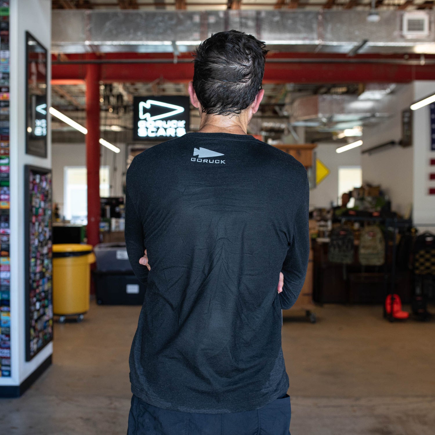 A man with wet hair stands indoors, facing away, wearing a black Men’s Embrace the Suck Long Sleeve - Tri-BlendX shirt in a gear shop.