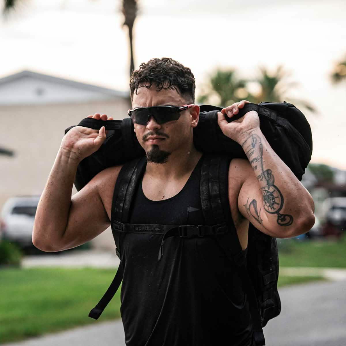 Athletic man wearing black performance tank and sunglasses carrying a heavy ruck sack outdoors