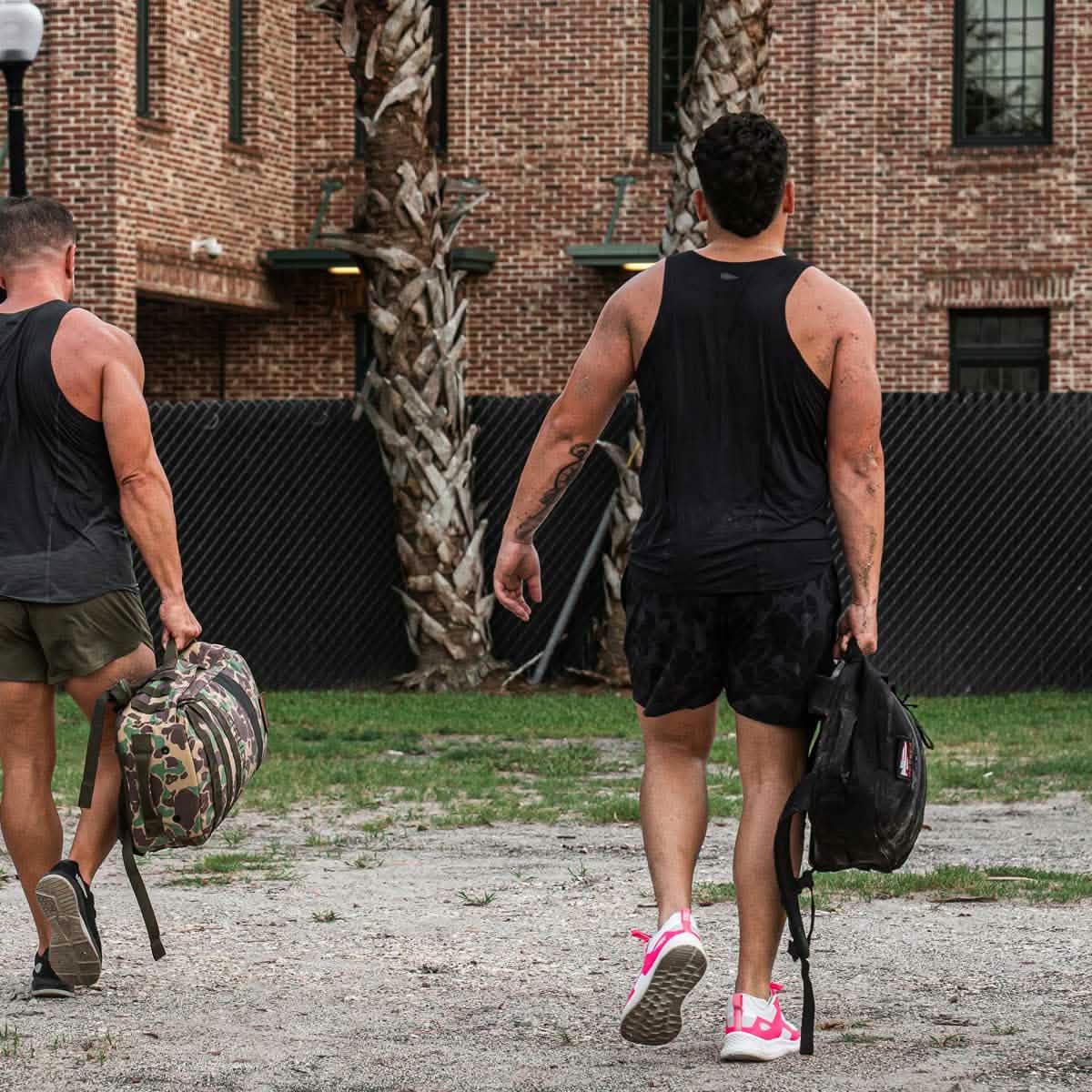 Two men wearing black performance tanks walking outdoors with outdoor bags near brick building and palm trees