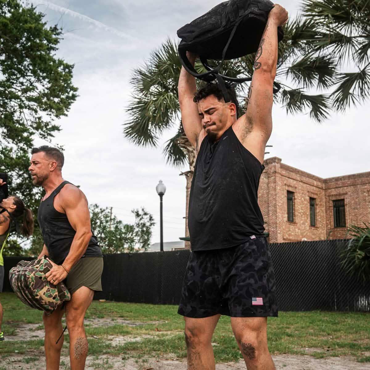 Two men in black performance tank tops lifting weighted sandbags outdoors during GORUCK training, with greenery and brick building in background