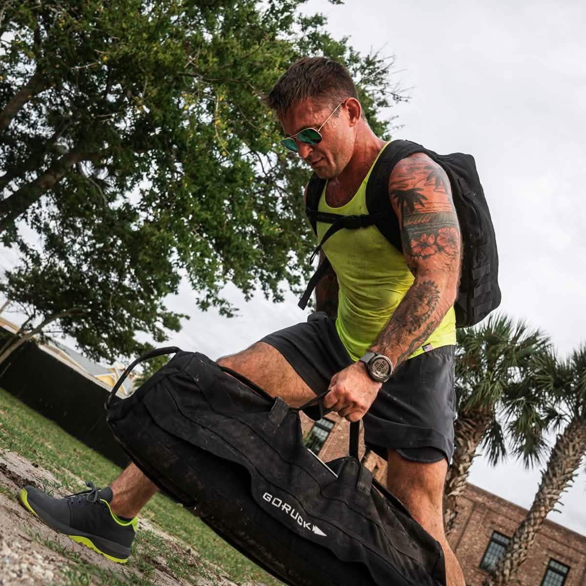 Athletic man wearing acid lime tank top and backpack lifting heavy black GORUCK bag outdoors