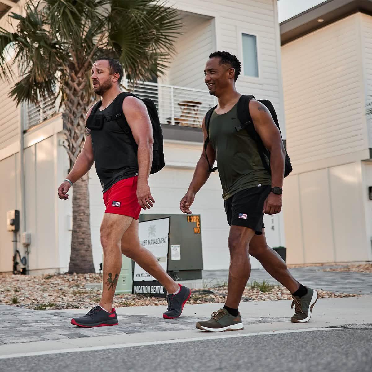 Two men rucking in GORUCK gear, wearing weighted backpacks, athletic shorts and tank tops, walking on a sunny neighborhood street.